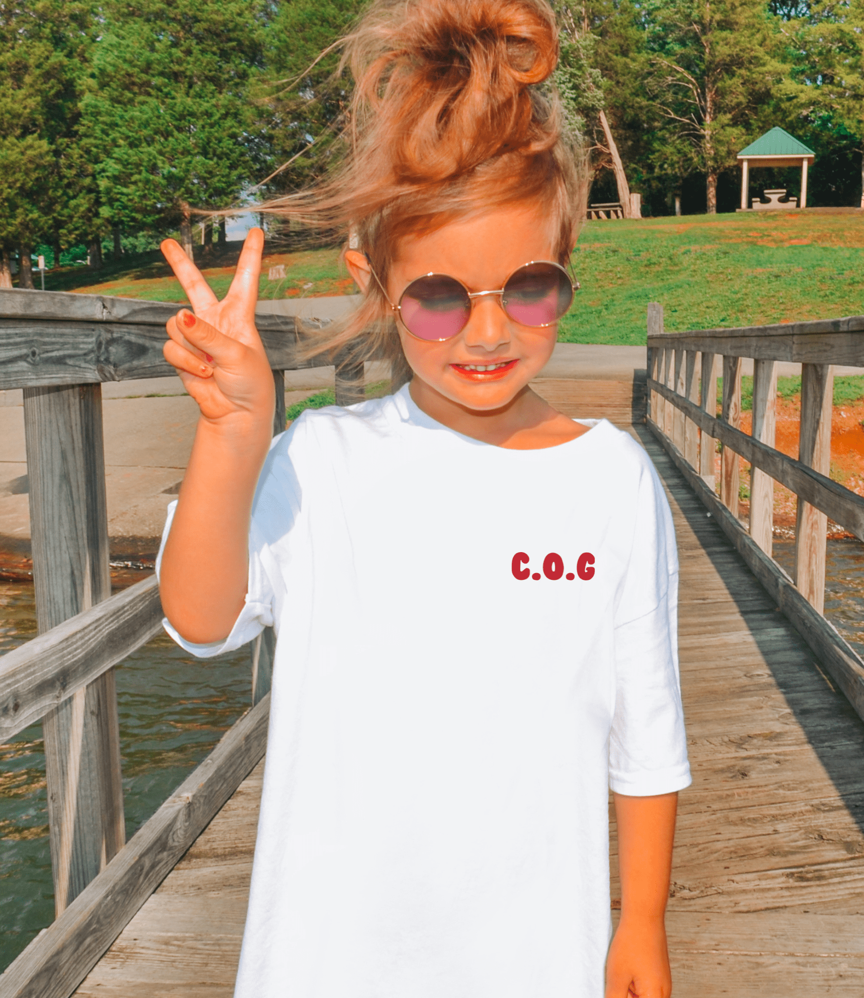 Child on a wooden bridge giving a peace sign, wearing a white shirt with 'C.O.G' in red pocket design. meaning Child of God