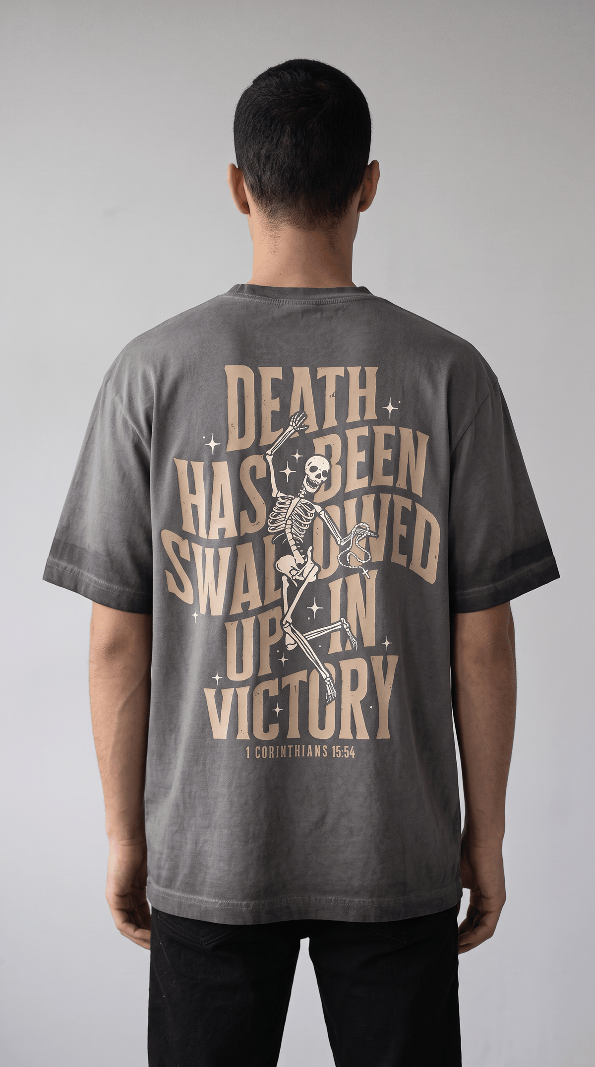 A man viewed from behind wearing a dark gray t-shirt with a large beige graphic print on the back featuring a skeleton and the text 'DEATH HAS BEEN SWALLOWED IN VICTORY'.