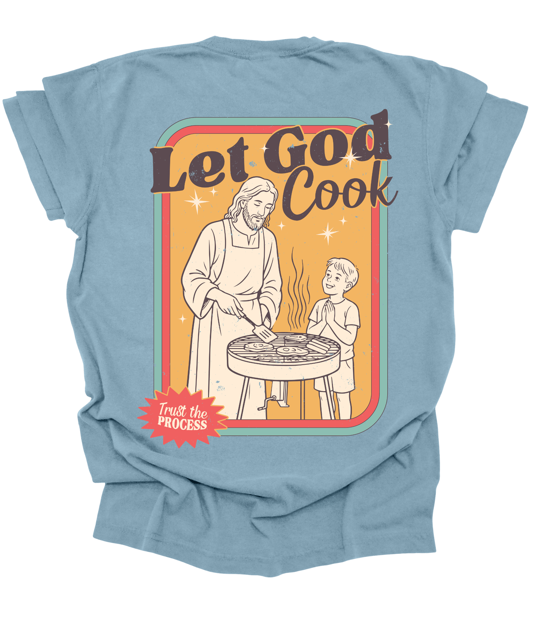 A light blue graphic t-shirt shown from the back, featuring a retro-style illustration of a man and a child grilling with the text 'Let God Cook' and a red starburst that reads 'Trust the Process'.
