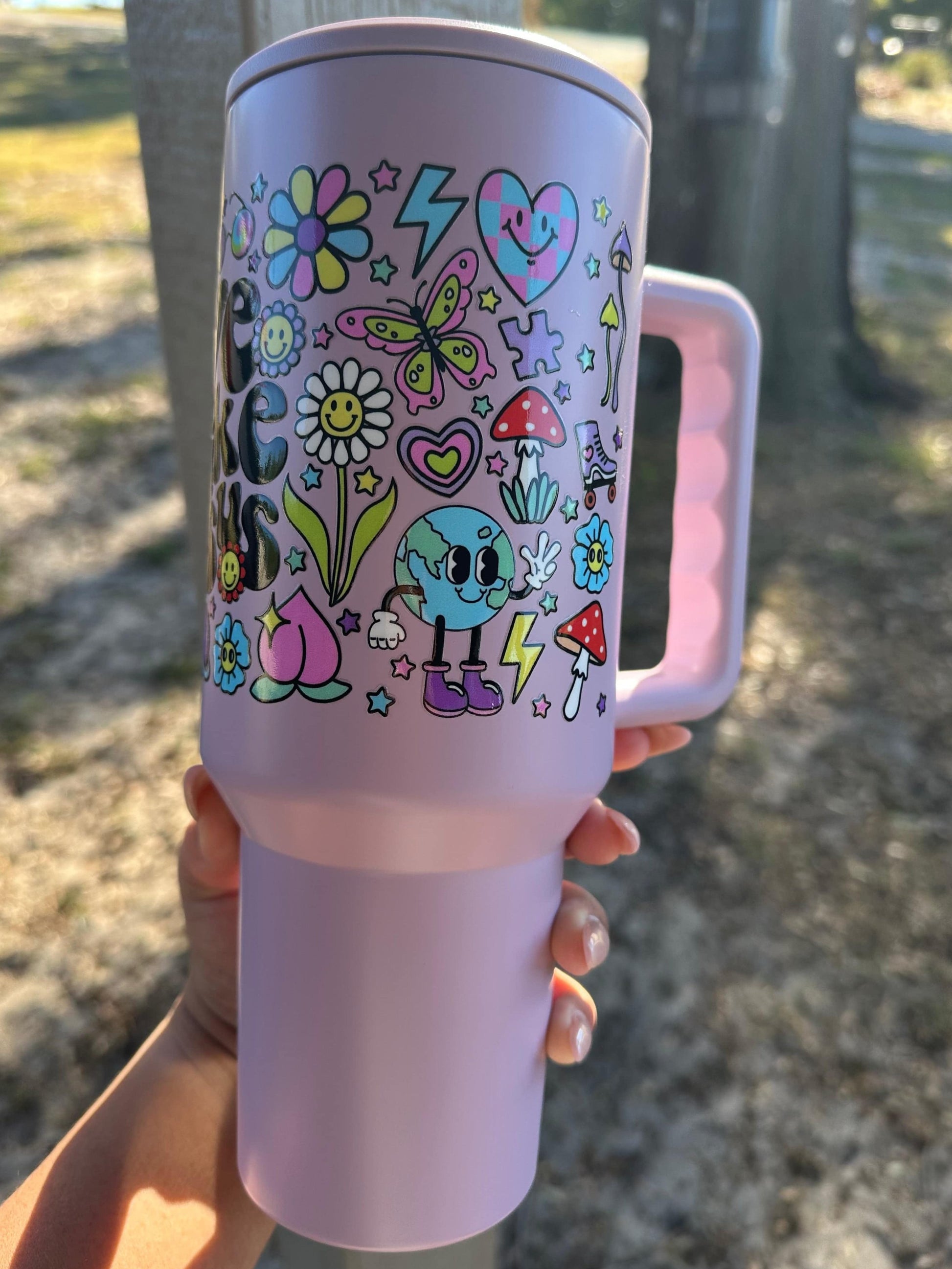 A lavender 40oz tumbler featuring colorful, faith-inspired designs and the phrase “Love Like Jesus” in bold lettering. The tumbler is held outdoors in natural light, showcasing vibrant details like rainbows, flowers, and smiley faces