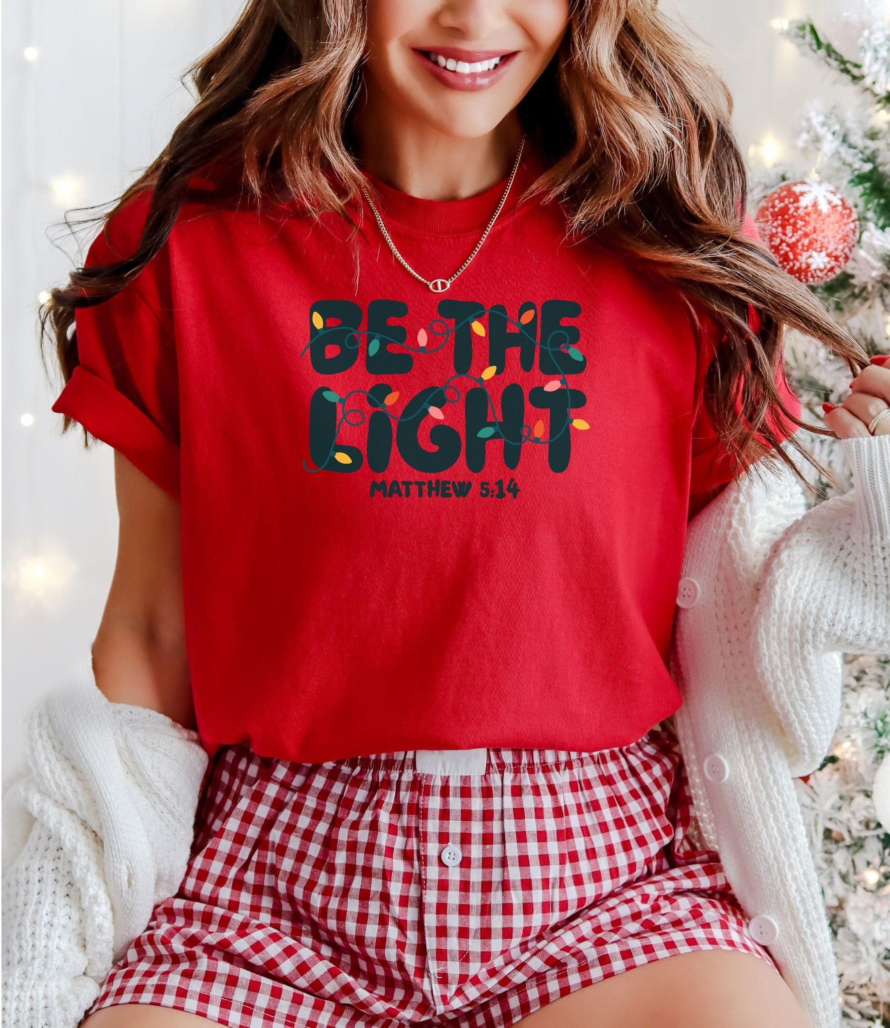 Person wearing a red shirt with 'Be the Light' text, sitting in a festive setting.