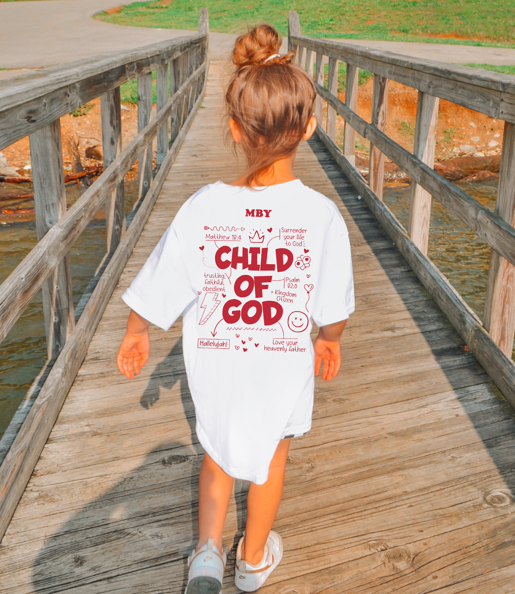 Child wearing a 'Child of God' t-shirt walking on a wooden bridge over water.