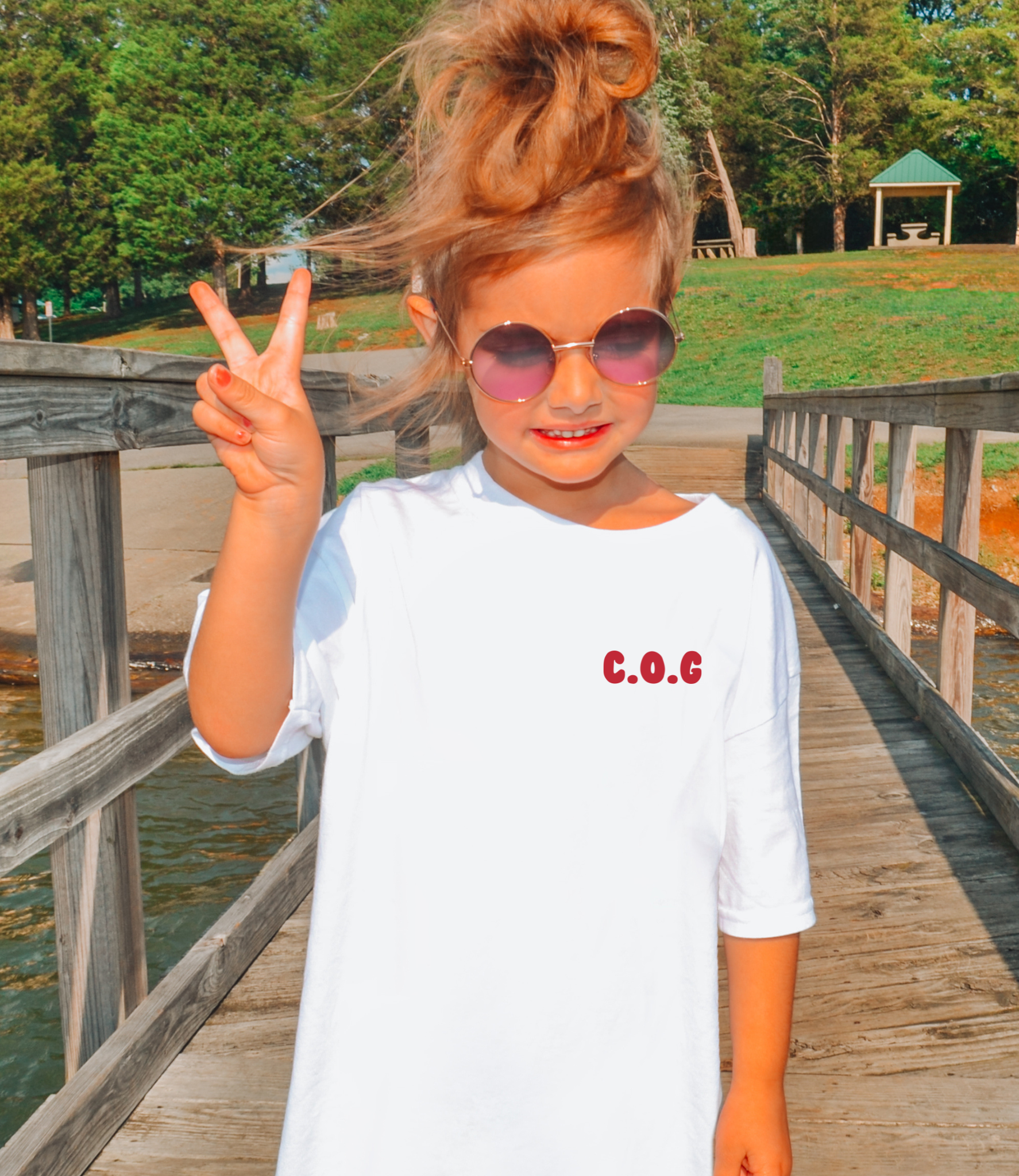 Child on a wooden bridge giving a peace sign, wearing a white shirt with 'C.O.G' in red pocket design. meaning Child of God