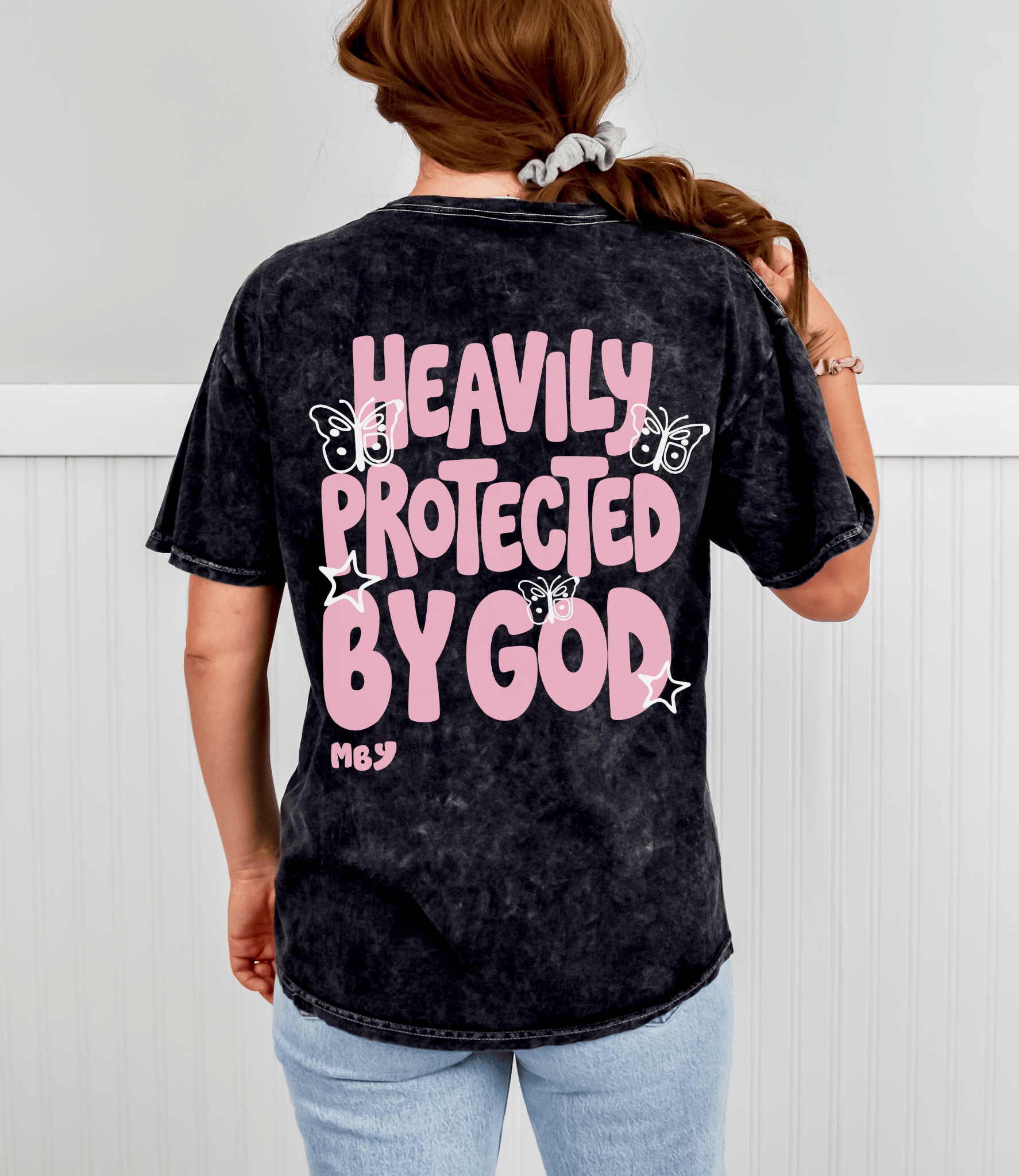 A woman viewed from behind wearing a black acid-wash t-shirt with a large pink and white graphic that reads 'HEAVILY PROTECTED BY GOD' with butterflies and stars, paired with light blue jeans.