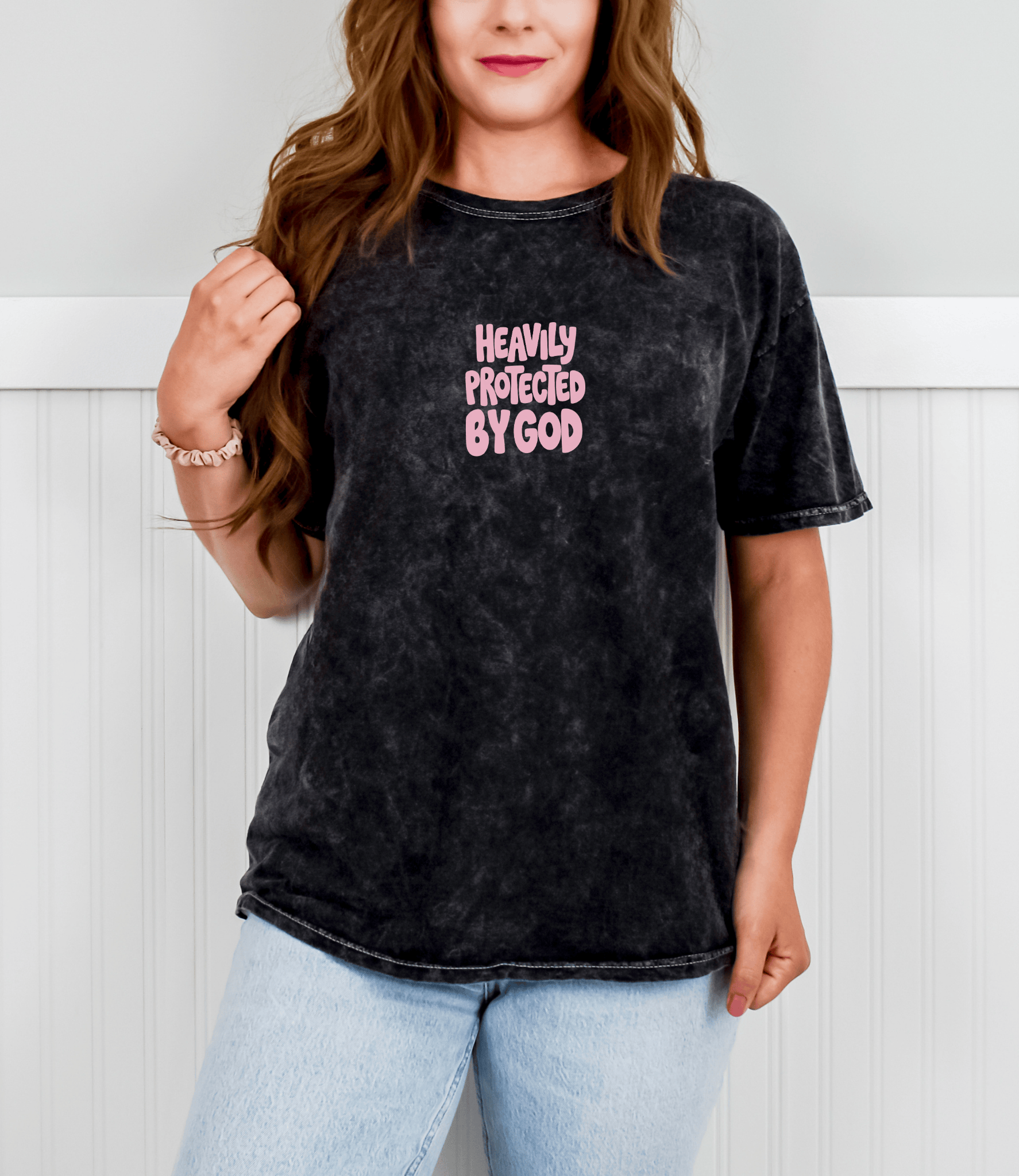 A woman wearing a black acid-wash graphic t-shirt with the text 'HEAVILY PROTECTED BY GOD' in pink, paired with light wash blue jeans, standing against a white paneled wall.