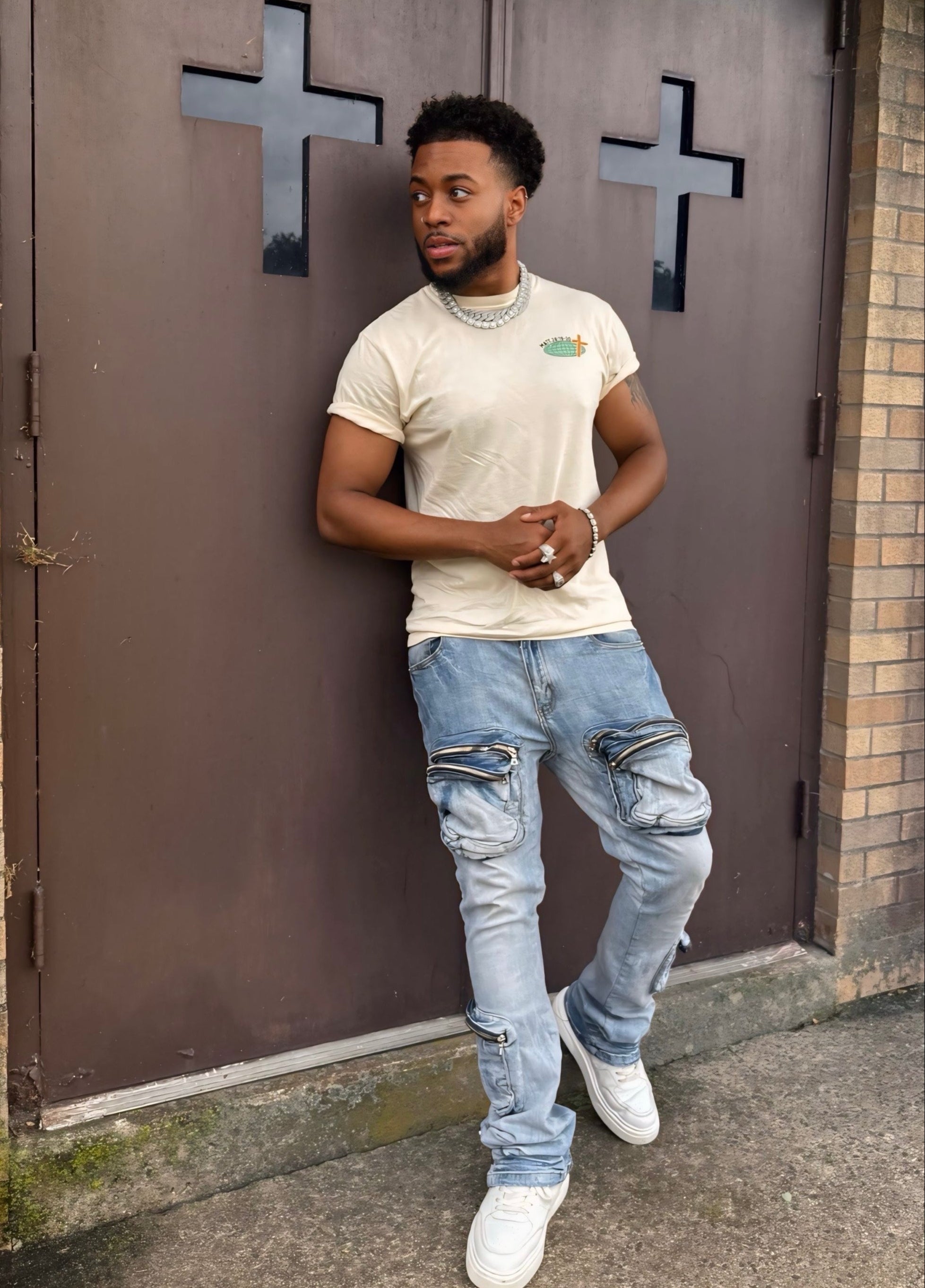Man leaning against a brown door with crosses, wearing a white t-shirt and light blue jeans.