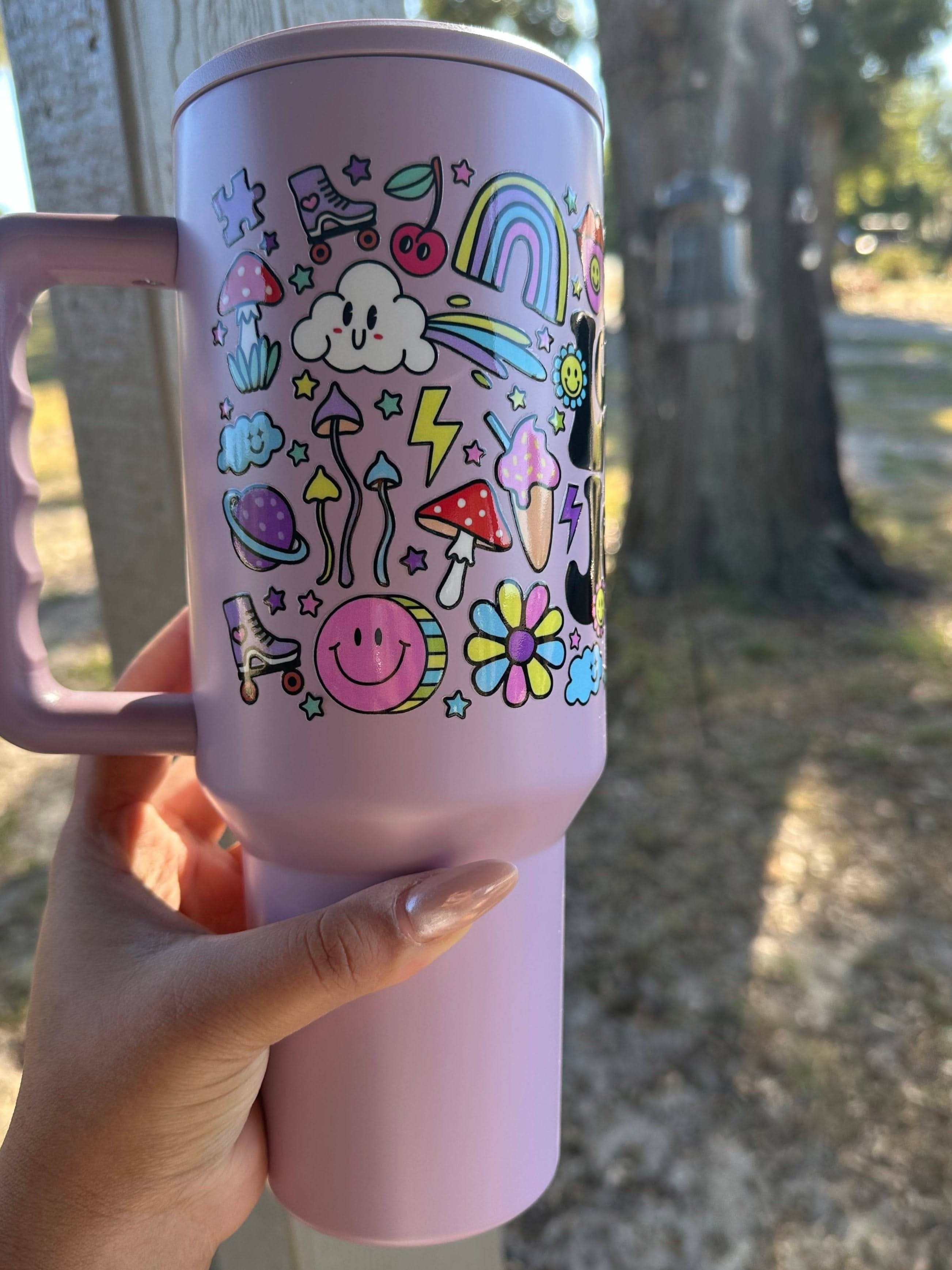 Side view of a lavender 40oz tumbler with a handle, displaying whimsical, multicolored illustrations such as rainbows, mushrooms, smiley faces, and stars. The photo captures the tumbler being held outside on a sunny day.